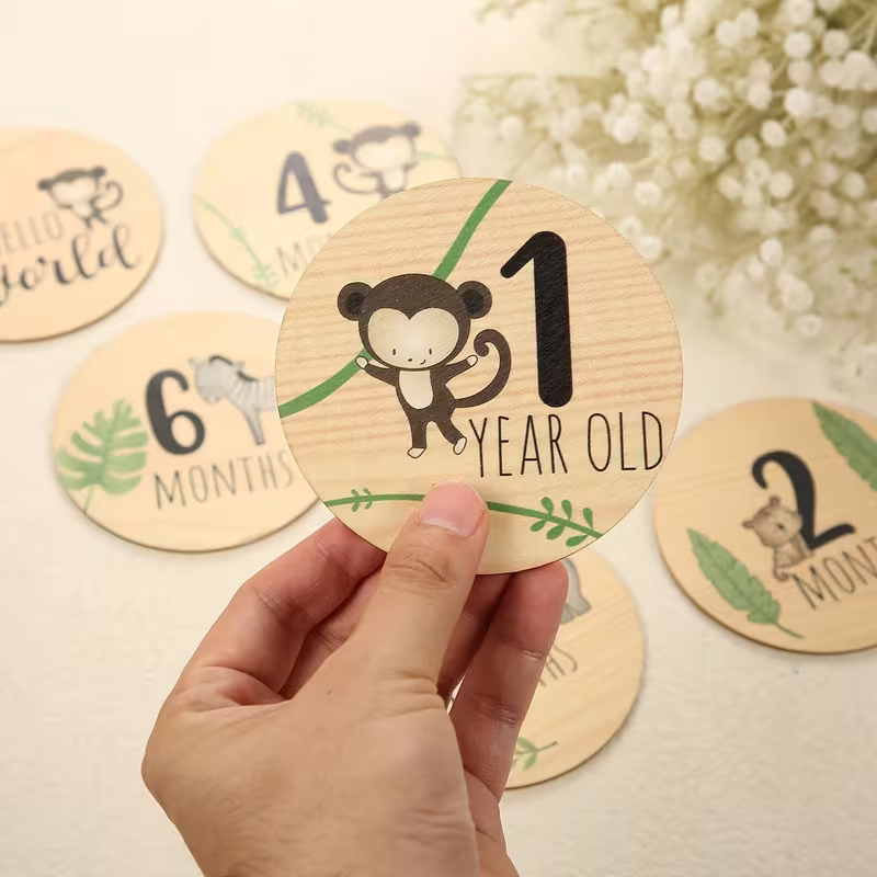 Hand holding a wooden milestone card with a monkey illustration and '1 year old' text, surrounded by other milestone cards on a light background.