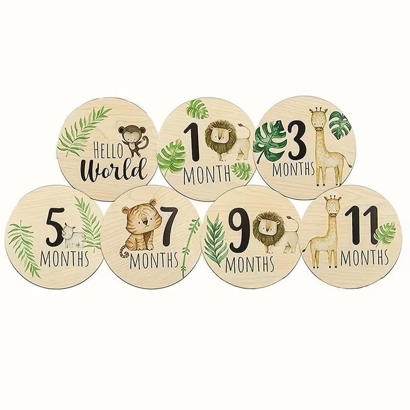 Set of seven round wooden baby milestone plaques with animal illustrations and text for 1, 3, 5, 7, 9, 11 months, and 'Hello World' on a light cream background.